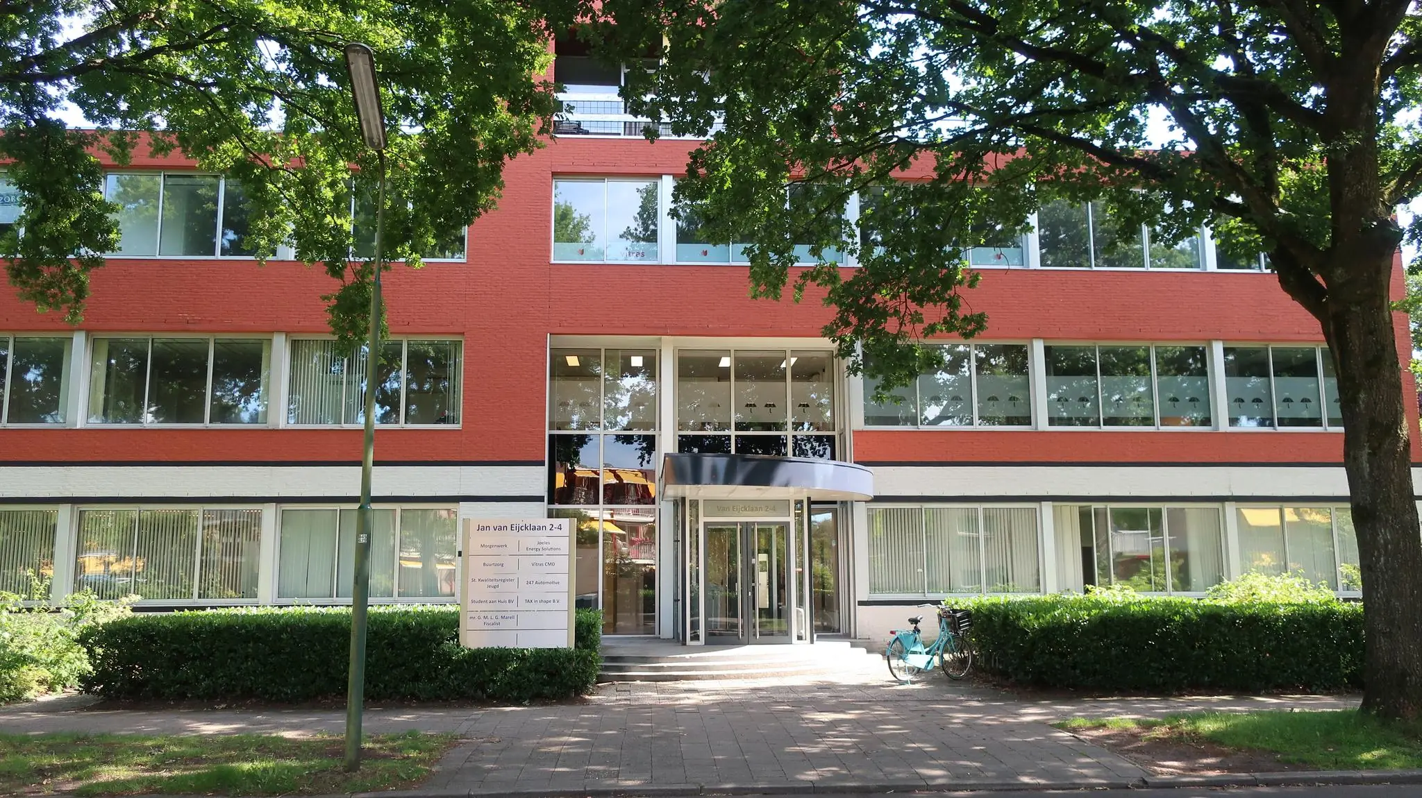 Vooraanzicht van een modern kantoorgebouw aan de Jan van Eycklaan 2-4, omgeven door bomen en een trottoir.